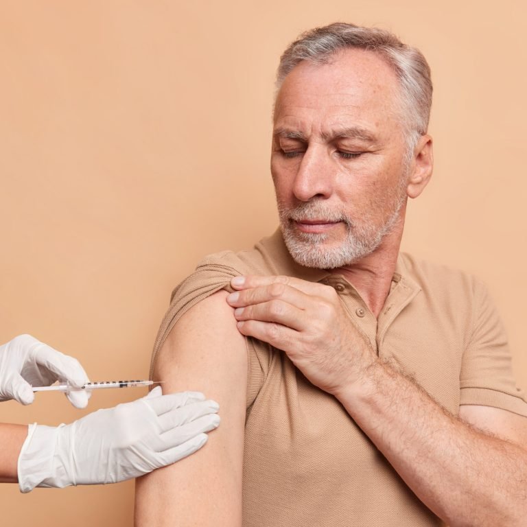 Serious grey haired old man gets vaccine injection against coronavirus wears t shirt isolated over beige studio background. Unrecognizable nurse vaccinates patient in clinic. Health care concept